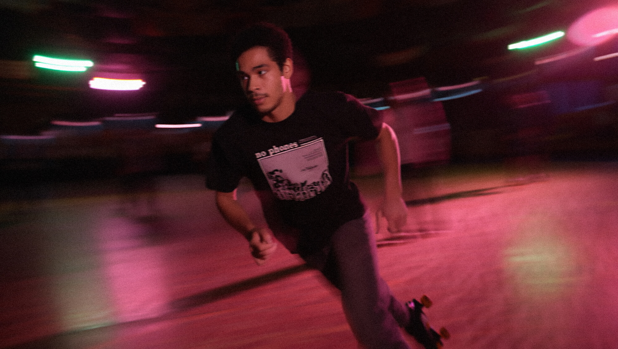 A roller skater goes around the rink with blurred neon lights and a t-shirt saying 'No phones'