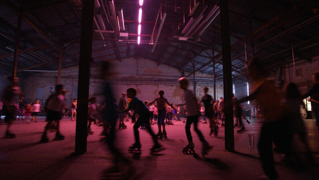 Hundreds of kids Roller Skating in an industrial roller skating rink under pink fluorescent light.