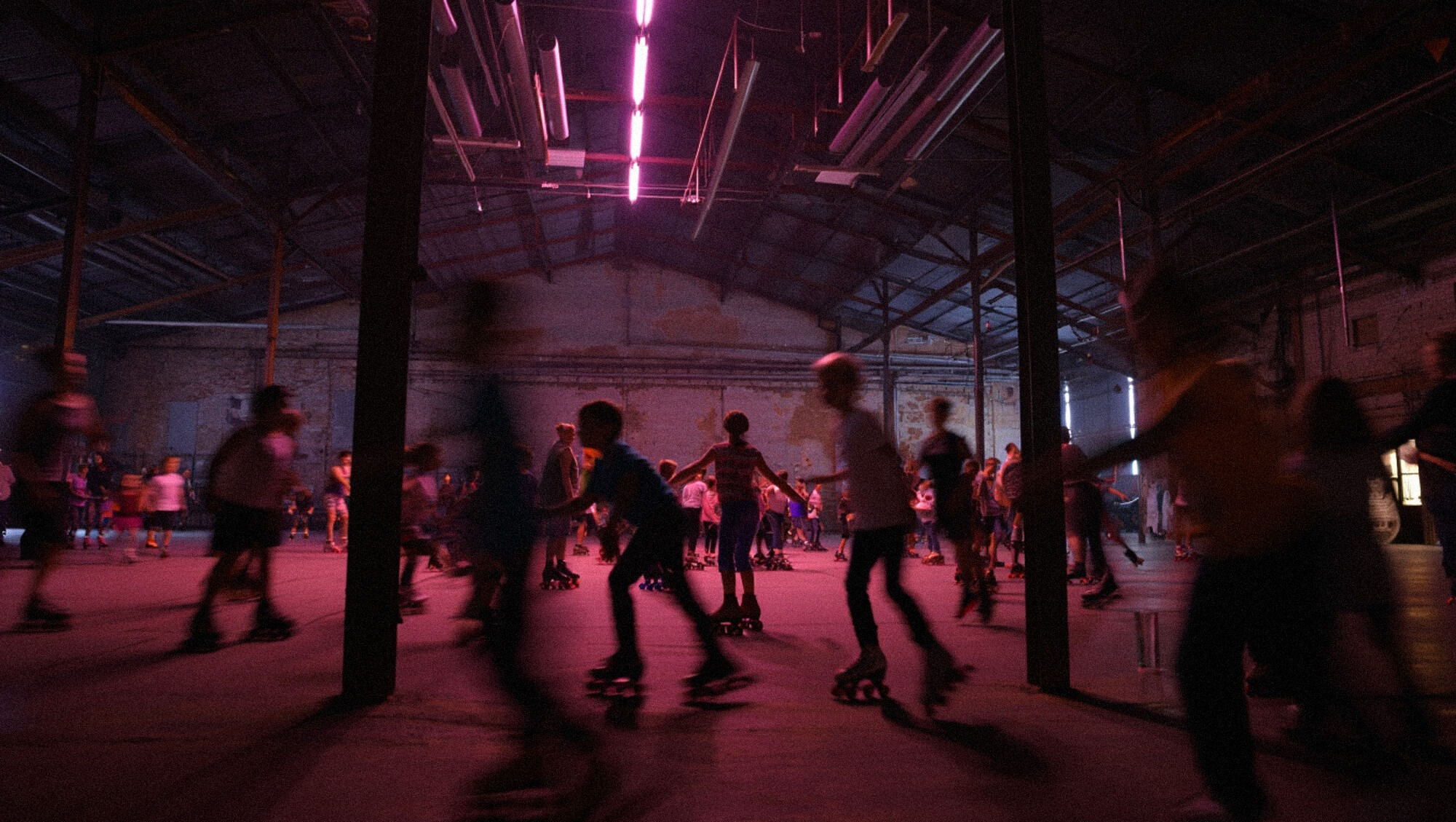 Hundreds of kids Roller Skating in an industrial roller skating rink under pink fluorescent light.